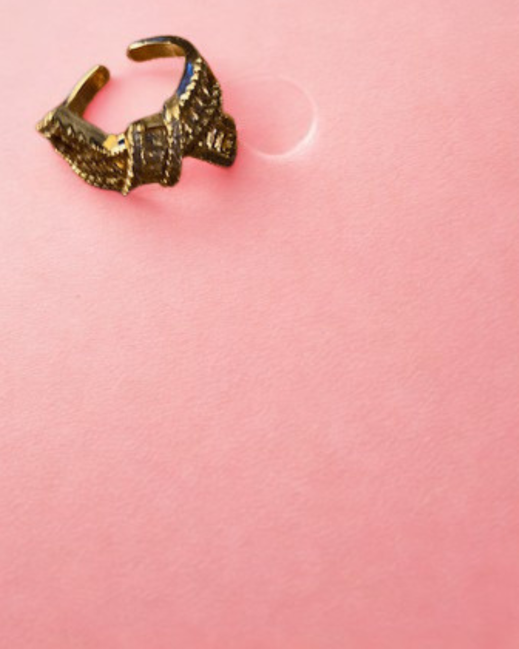 A textured gold-toned ring with a geometric, woven pattern, photographed on a solid pink background.