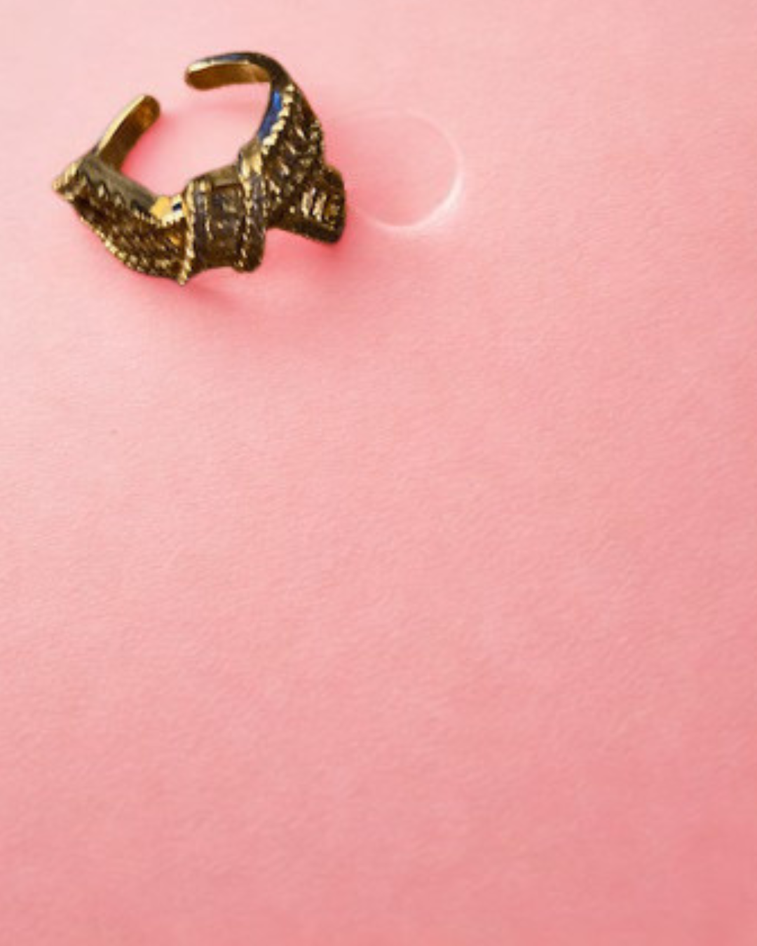 A textured gold-toned ring with a geometric, woven pattern, photographed on a solid pink background.